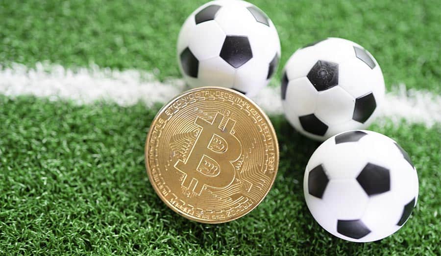 Três mini bolas de futebol ao lado de uma moeda de Bitcoin.