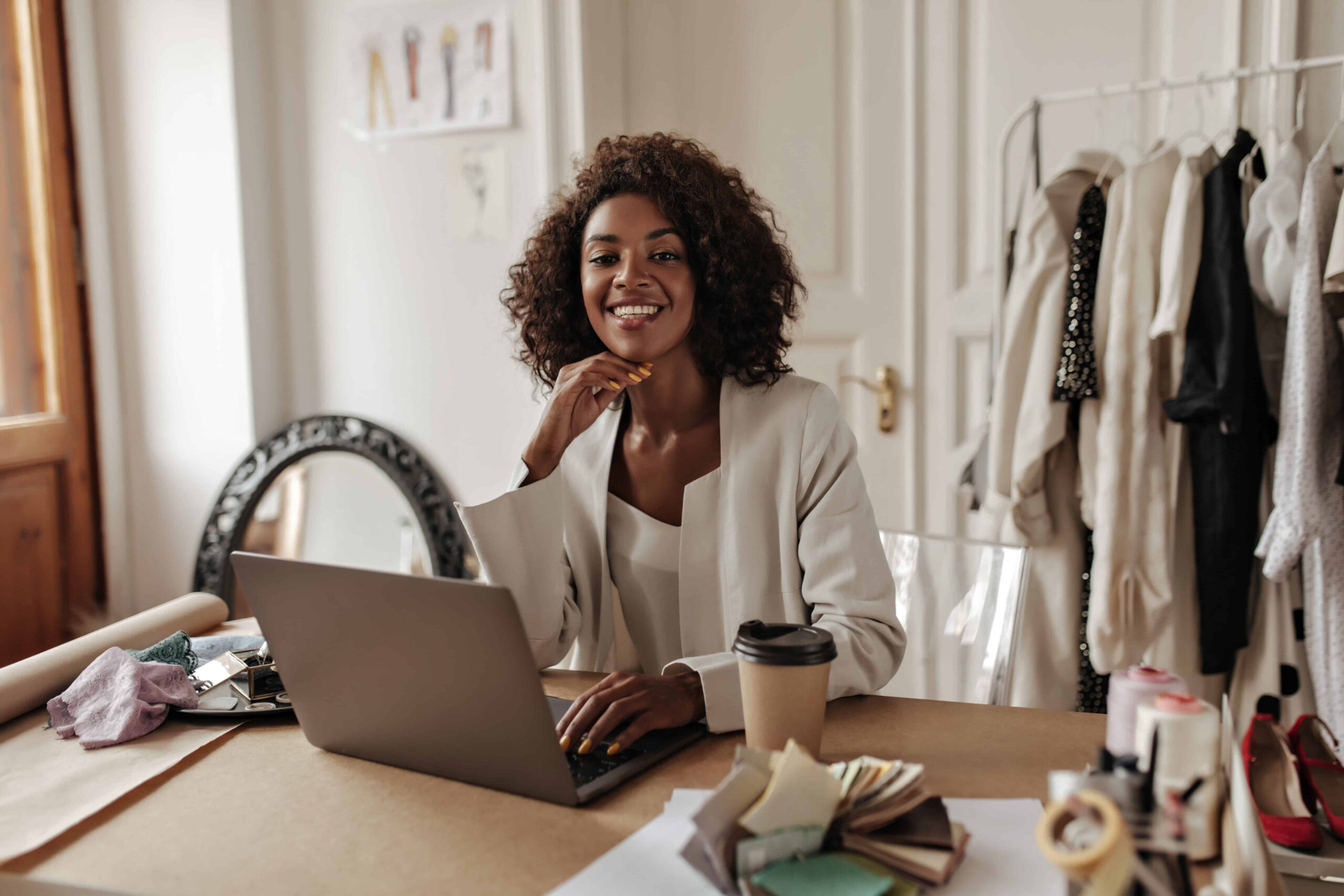 charming young darkskinned woman in stylish jacket and blouse smiles looks at camera works in laptop and poses in office scaled