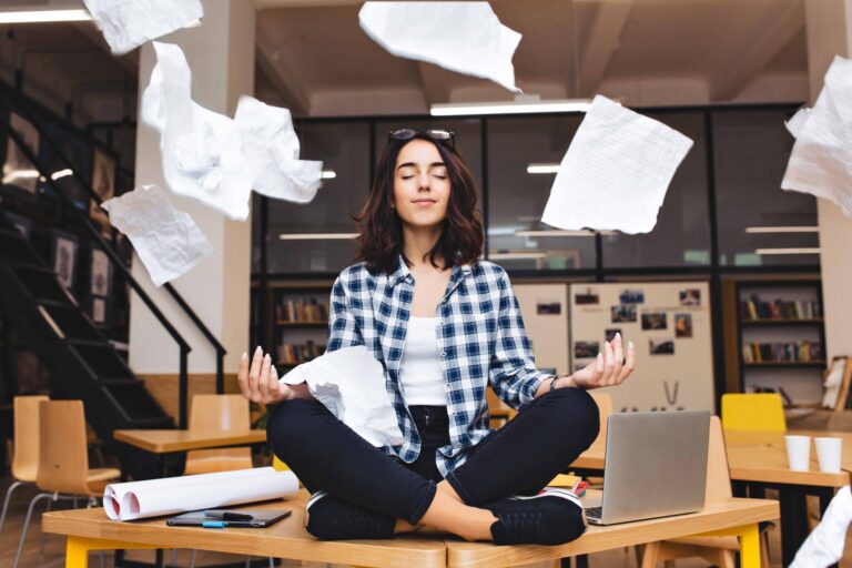young pretty joyful brunette woman meditating on table surround work stuff and flying papers cheerful mood taking a break working studying relaxation true emotions scaled