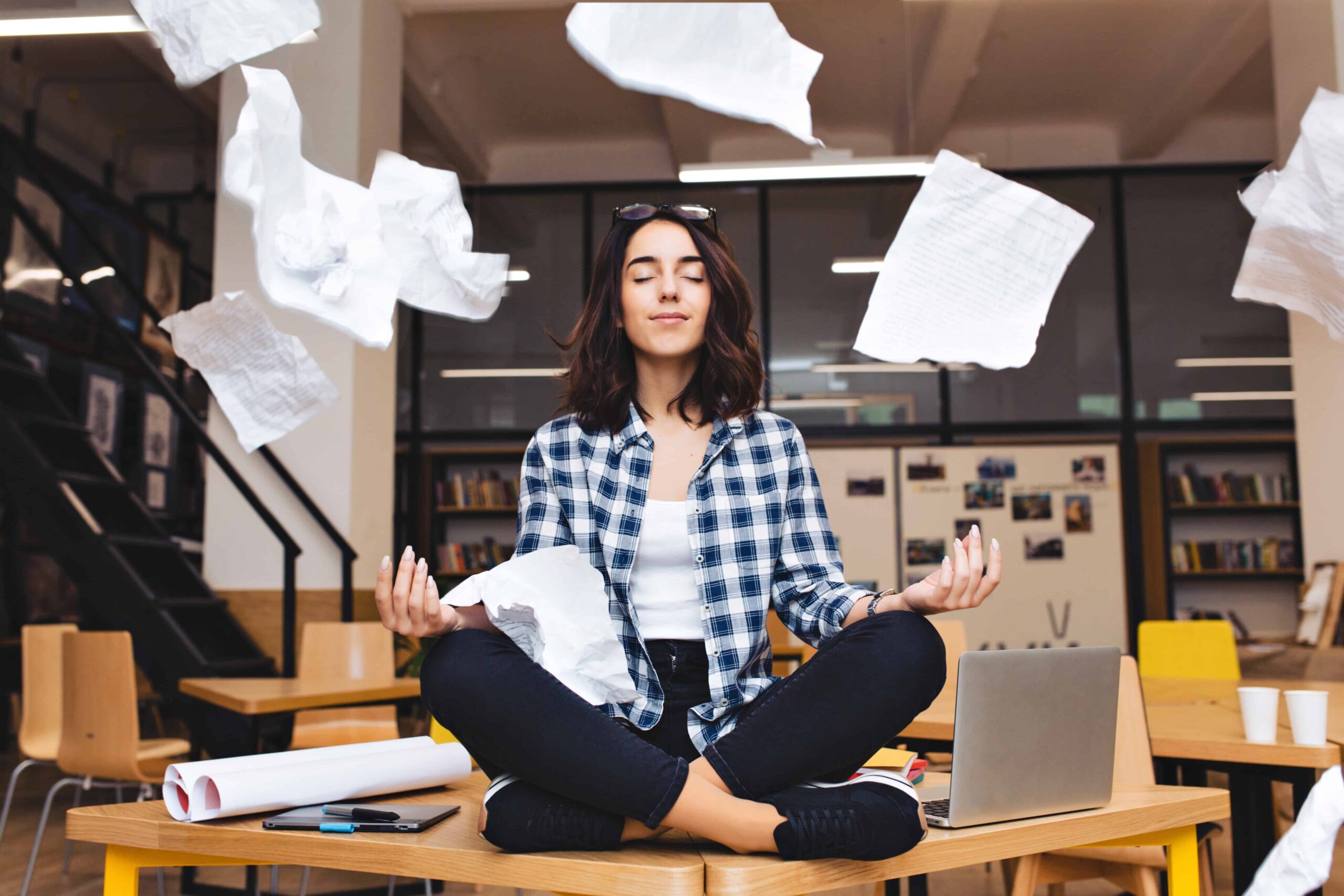 young pretty joyful brunette woman meditating on table surround work stuff and flying papers cheerful mood taking a break working studying relaxation true emotions scaled