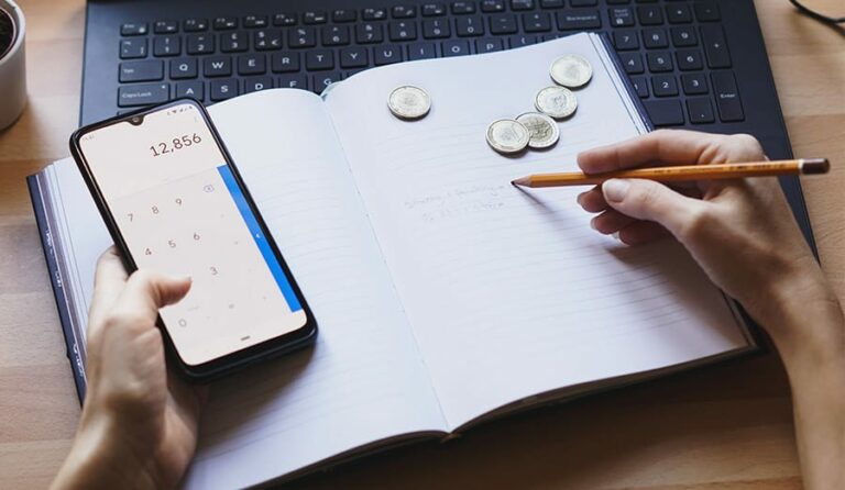 Pessoa com celular e caderno na mão calculando gastos.
